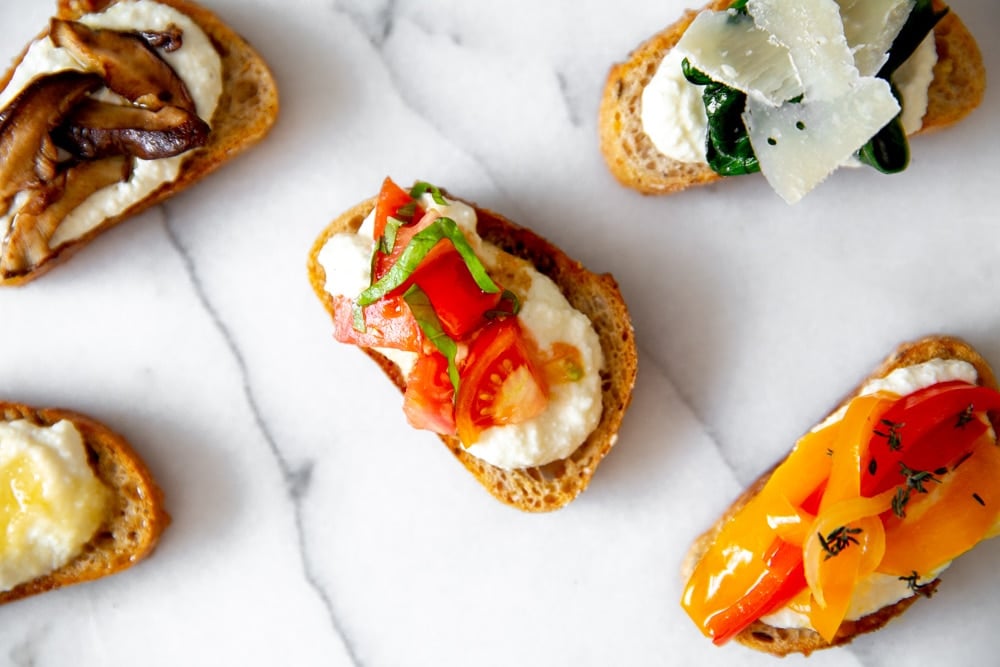 Overhead shot showing five different ricotta crostini on a marble surface.