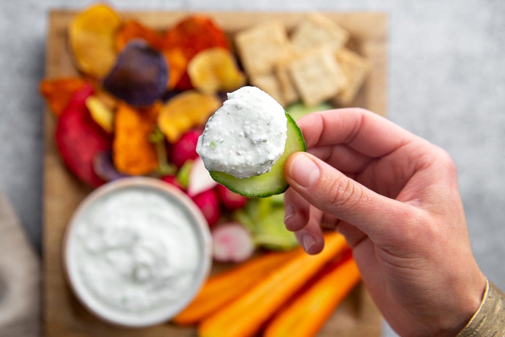 Close up of a hand holding a cucumber topped with the herbed cottage cheese dip.