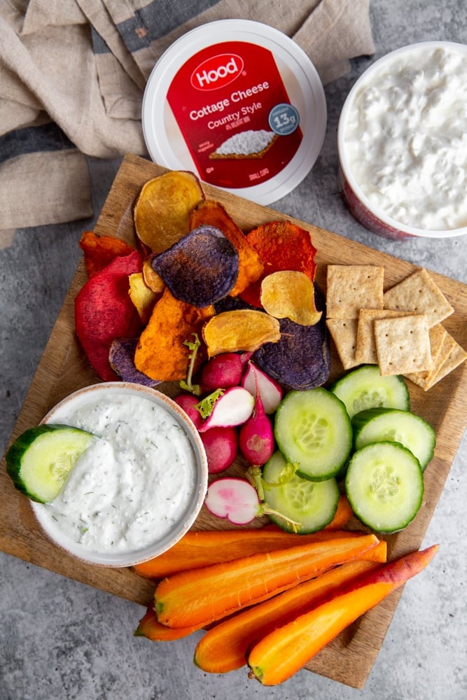 The creamy veggie dip on a serving board with sliced veggies and crackers, with a container of cottage cheese alongside.