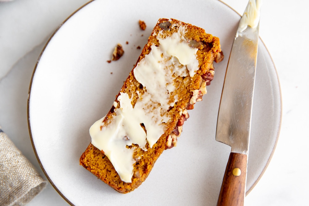 Close up of a slice of buttered pumpkin bread on a plate with a knife alongside.