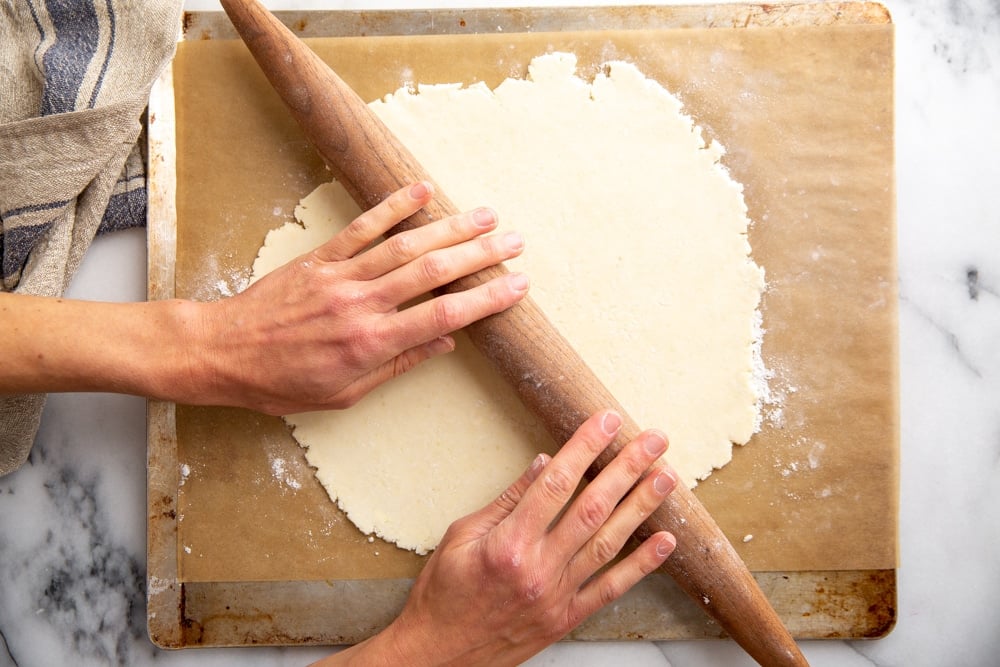 Hands rolling out pie dough on a piece of parchment paper. 