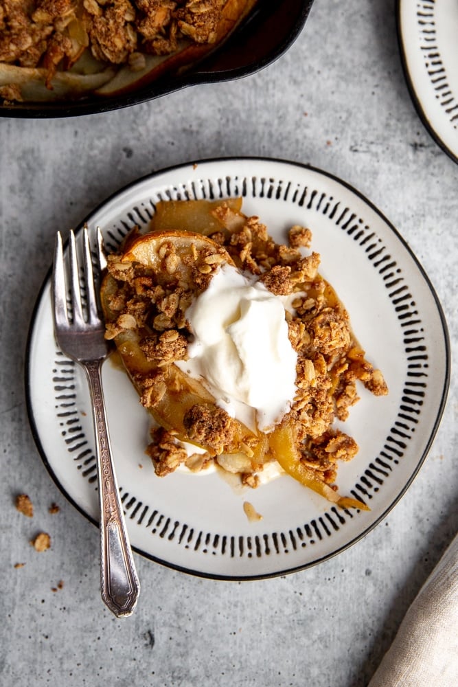 Close up of a serving of pear crumble on a plate topped with whipped cream, with a fork alongside. 