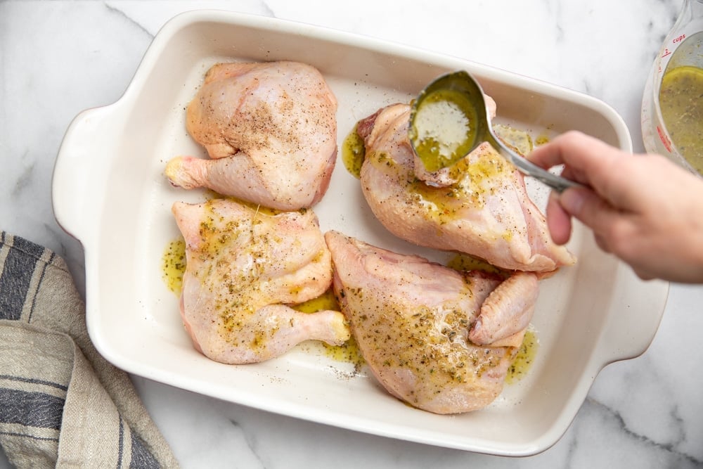 Process shot showing a hand drizzling the lemon marinade over chicken pieces in a baking dish. 