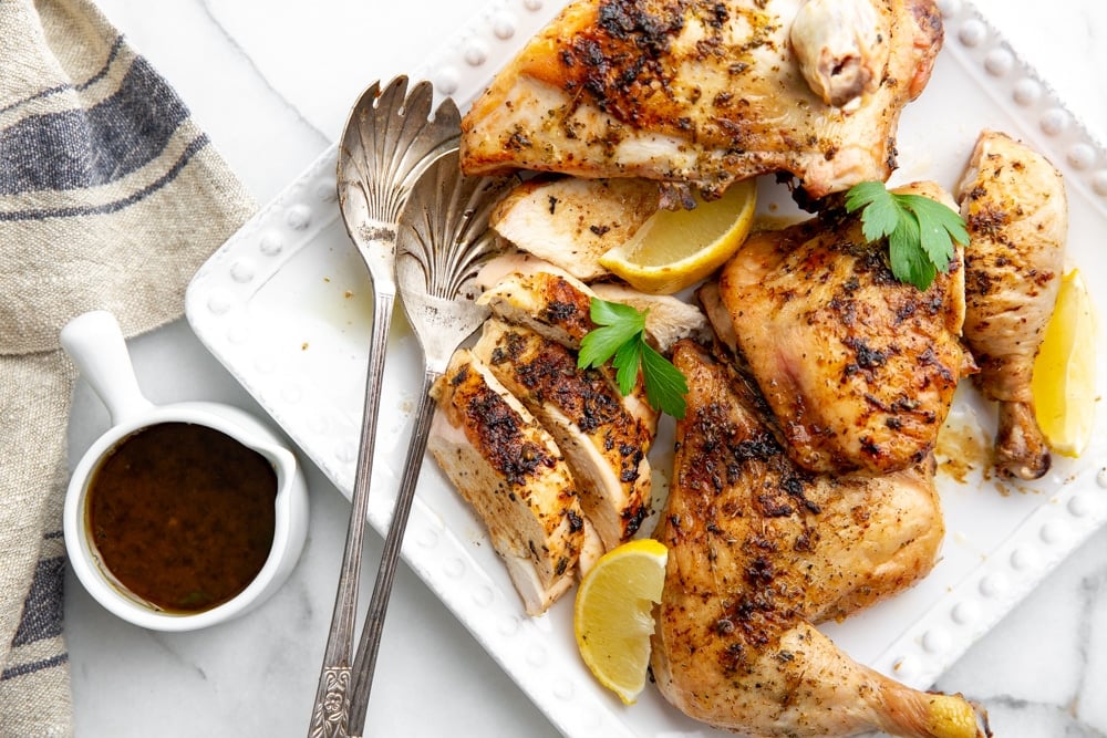 Roasted lemon garlic chicken on a serving platter with serving spoons, with the pan sauce alongside. 