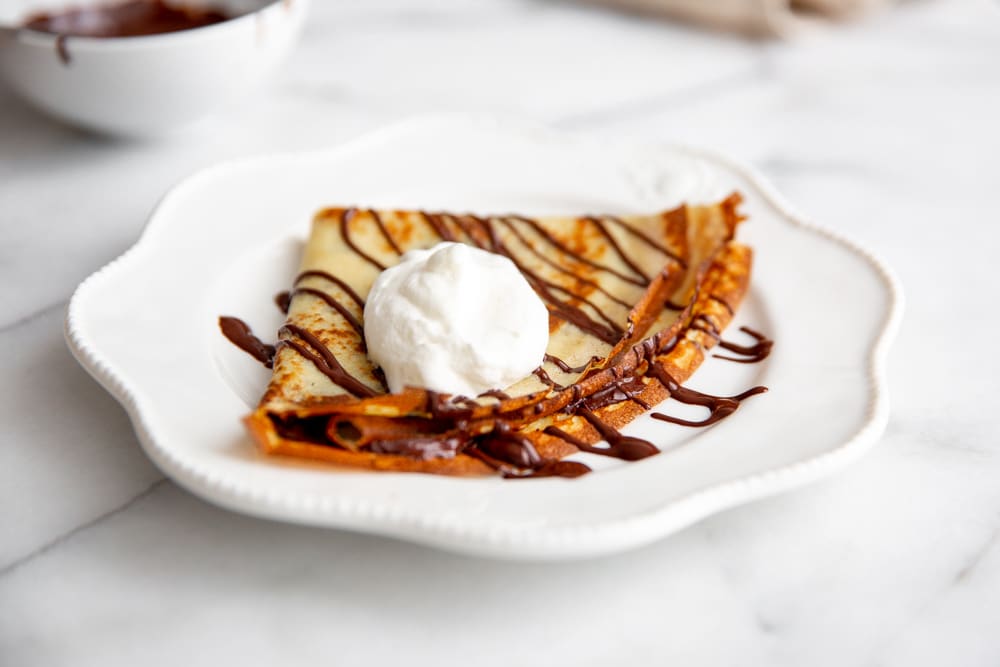 A chocolate crepe on a plate, drizzled with melted chocolate and topped with whipped cream. 