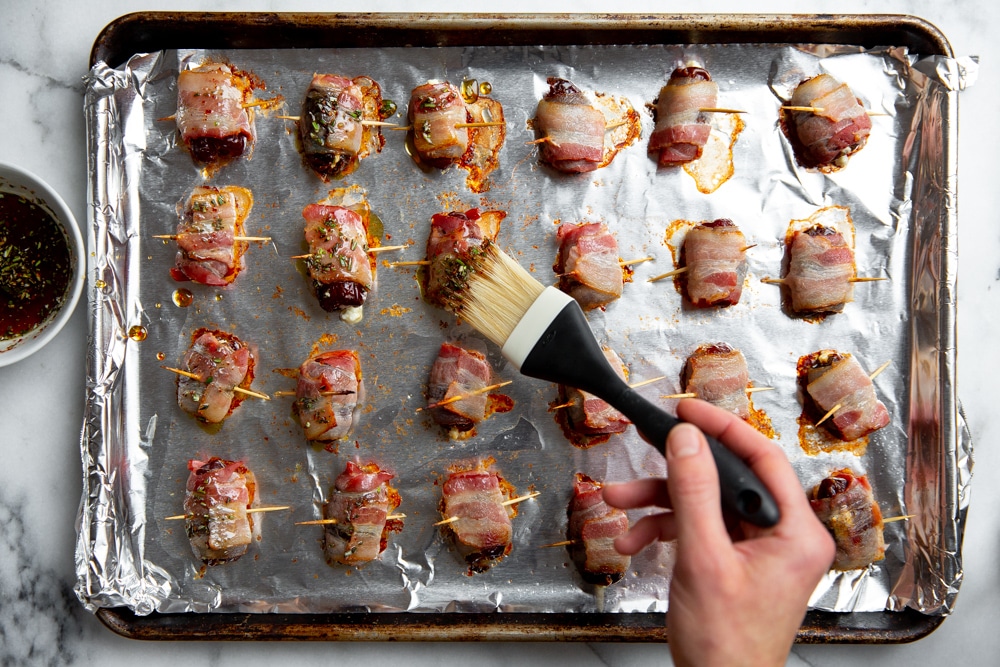 Process shot showing a hand brushing the bacon wrapped dates on a baking sheet with the maple glaze.