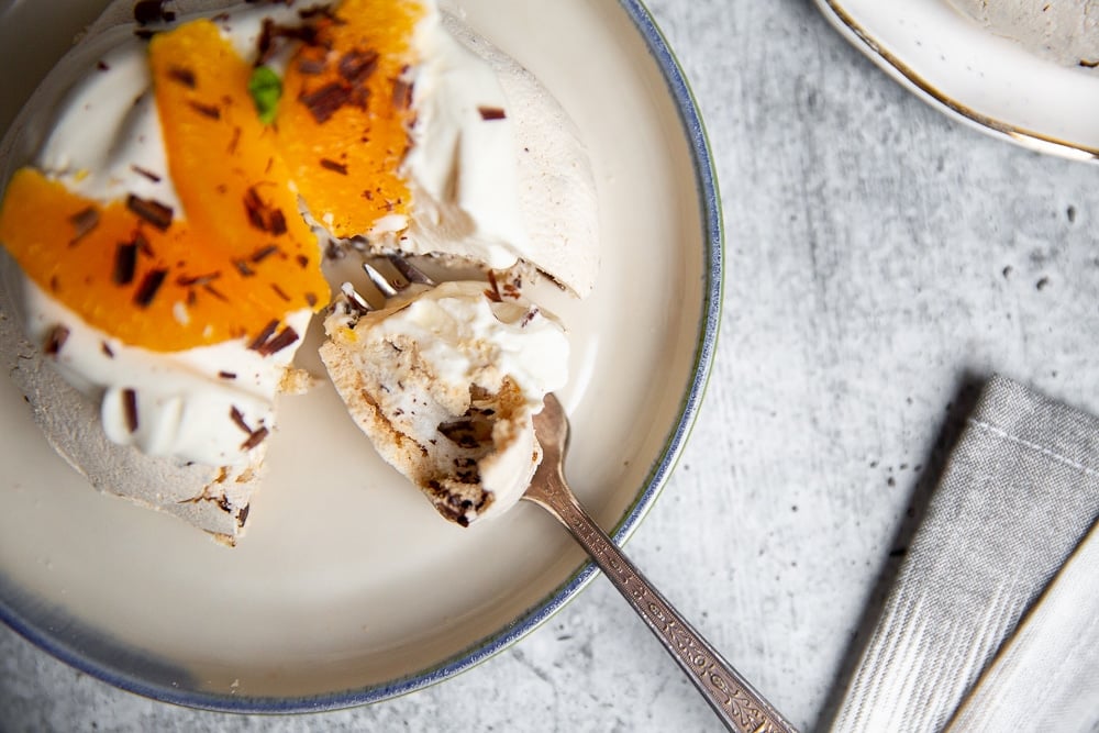 Pavlova on a plate topped with whipped cream and orange segments, with a fork taking a bite.