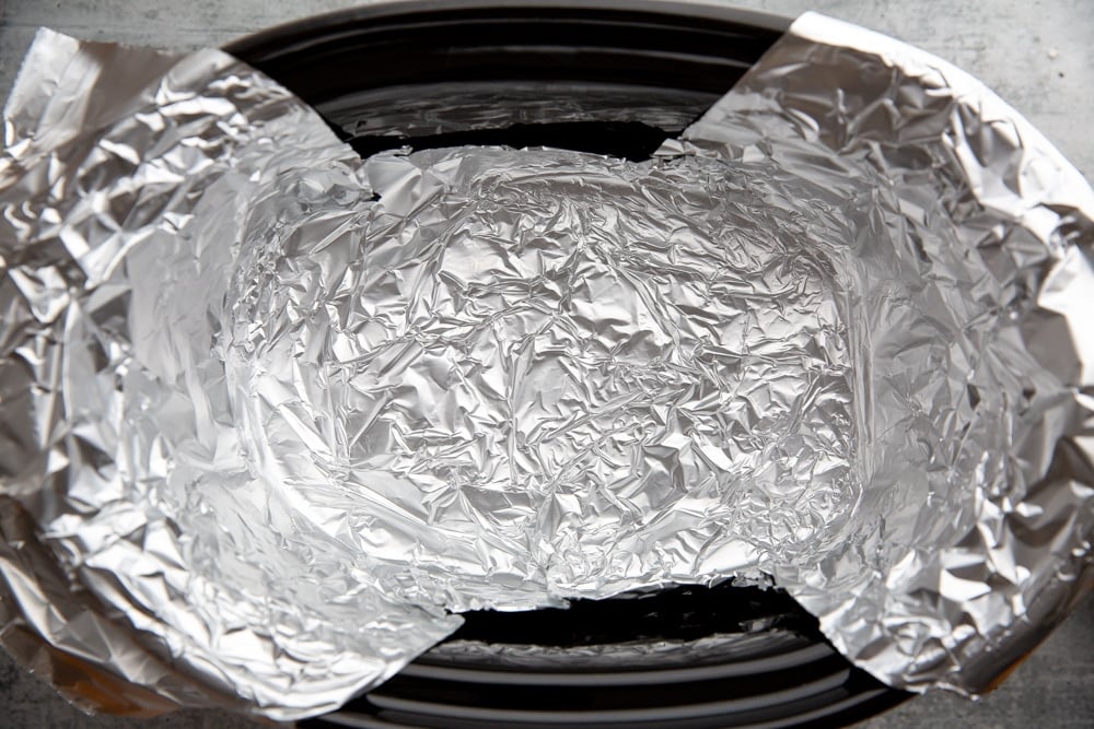 Process shot showing a slow cooker lined with aluminum foil. 