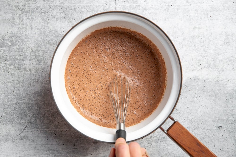 Process shot showing a hand whisking milk into the cocoa powder mix in a saucepan. 