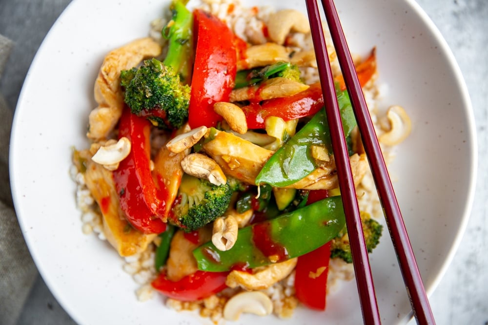 Close up of a stir fry in a bowl over rice with chopsticks.