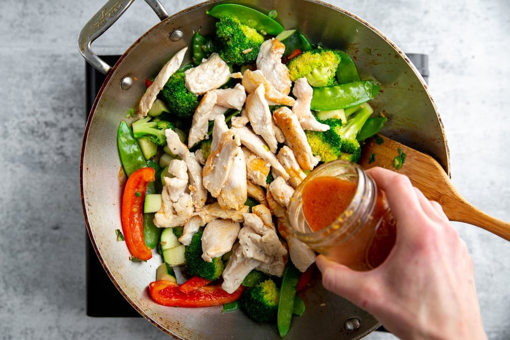 A hand pouring stir fry sauce into sauteed vegetables and chicken in a wok.