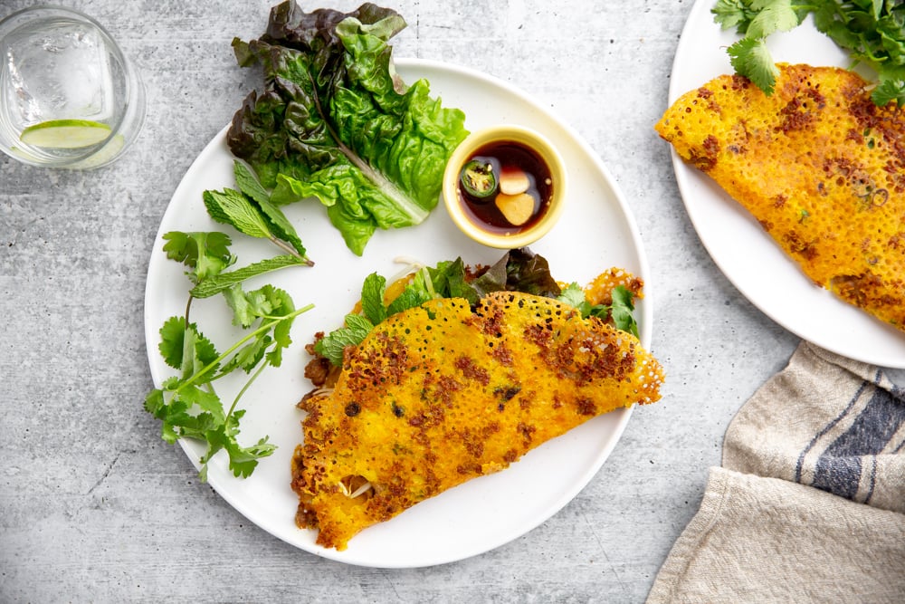 Vietnamese rice flour crepes on a plate with lettuce leaves, cilantro, mint and a dipping sauce.