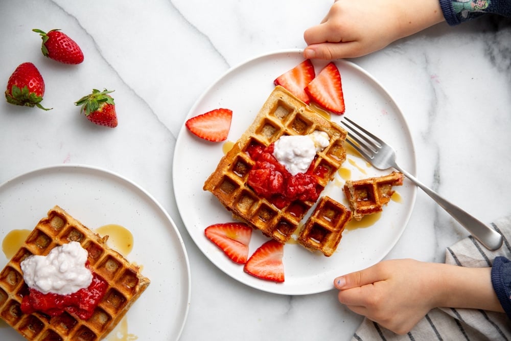 Two child hands grabbing a plate of waffles topped with strawberries and cottage cheese.