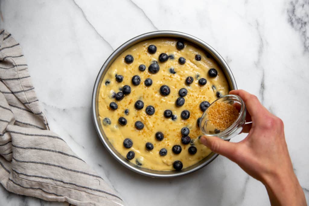 Hand sprinkling turbinado sugar over the blueberry cake batter in a cake pan.
