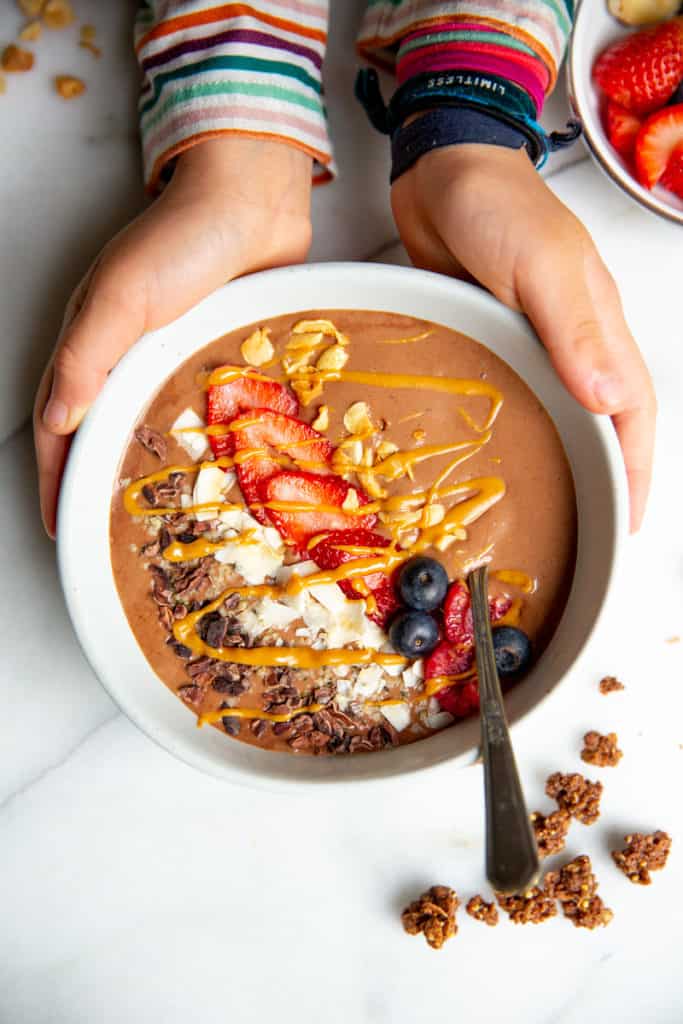 Kids hands wrapped around a chocolate peanut butter banana smoothie bowl. 