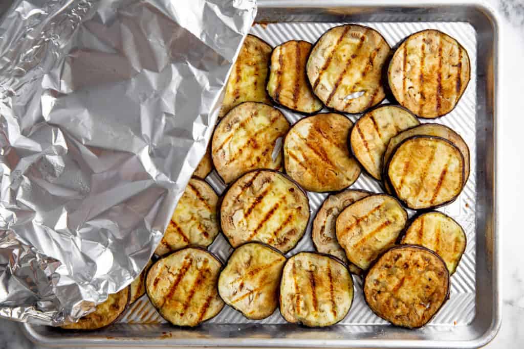 Process shot showing eggplant slices covered with foil on a sheet pan. 