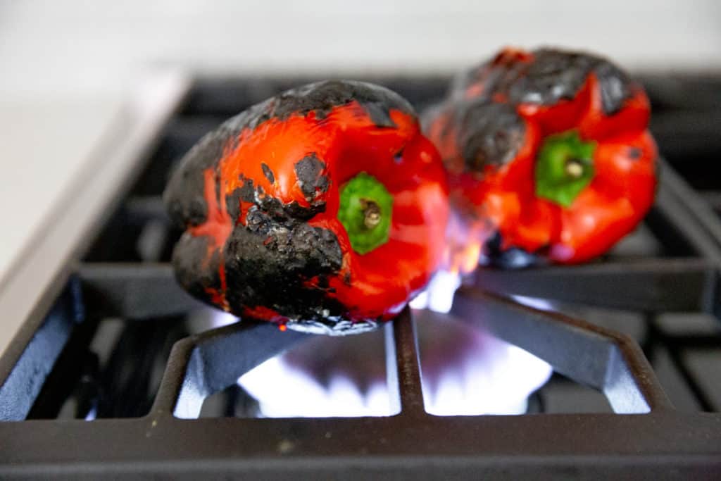 Process shot showing red peppers roasting over a gas flame on a stovetop.