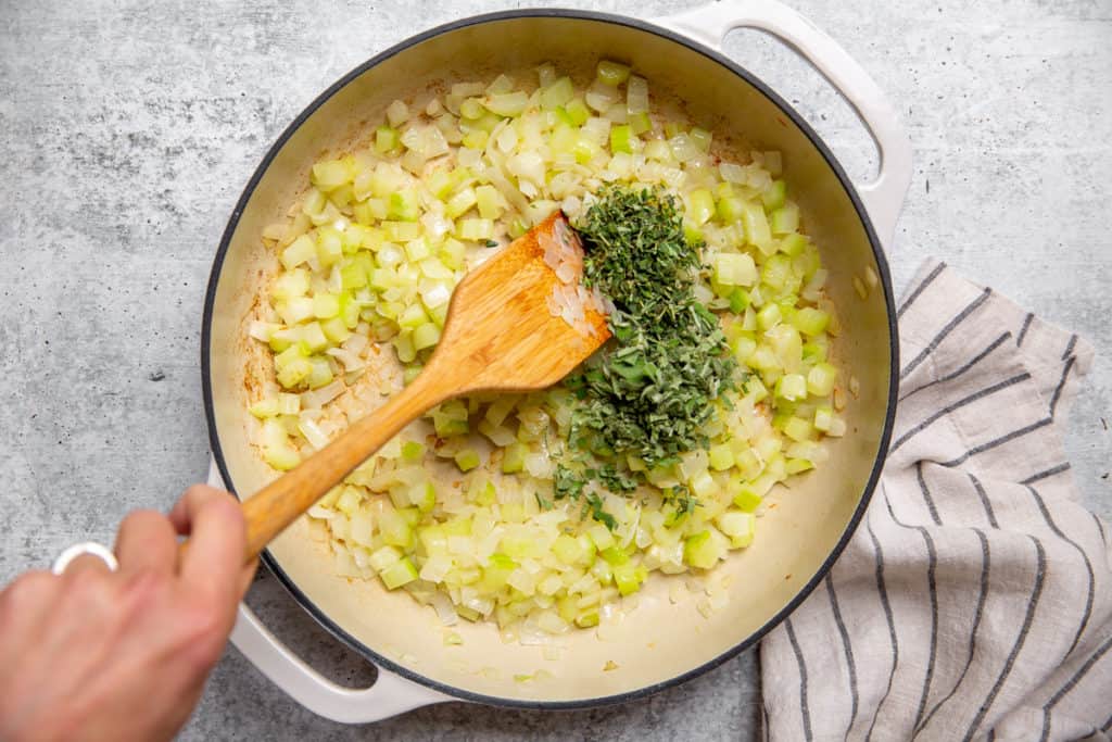 Wooden spoon stirring chopped sage and thyme into a skillet with cooked onion and celery. 