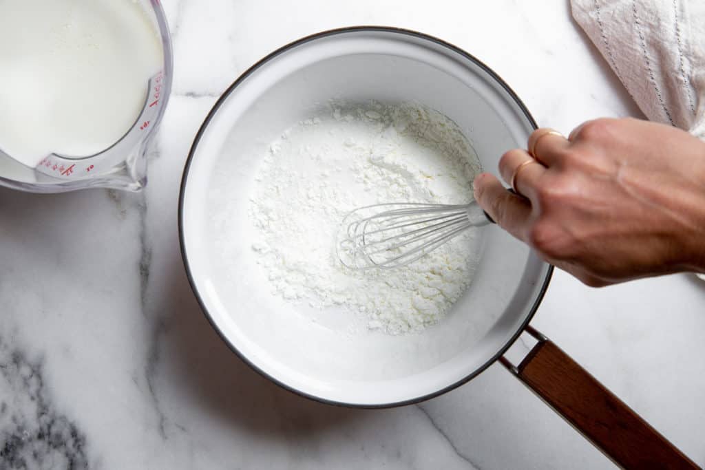 Hand whisking sugar, cornstarch and salt in a saucepan, with milk alongside.