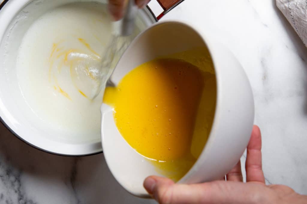 Hand pouring the tempered egg mixture back into the pudding in the saucepan. 