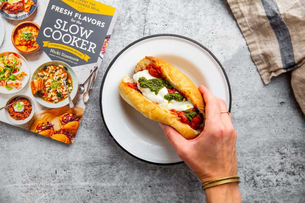 A hand holding a meatball sub with the book, Fresh Flavors for the Slow Cooker alongside.