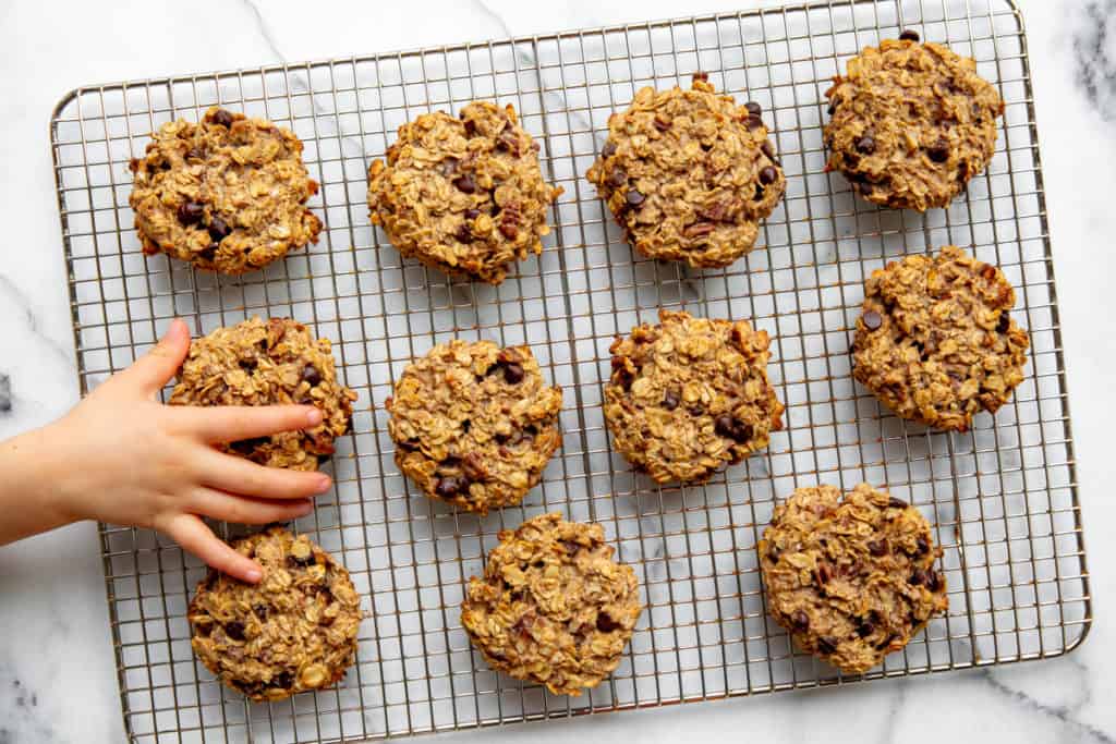 A kid's hand grabbing a banana oatcake cookie off of a cooling rack. 