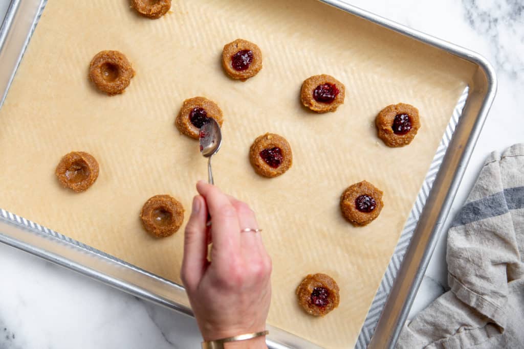 A hand spooning jam into the thumbprint cookies. 