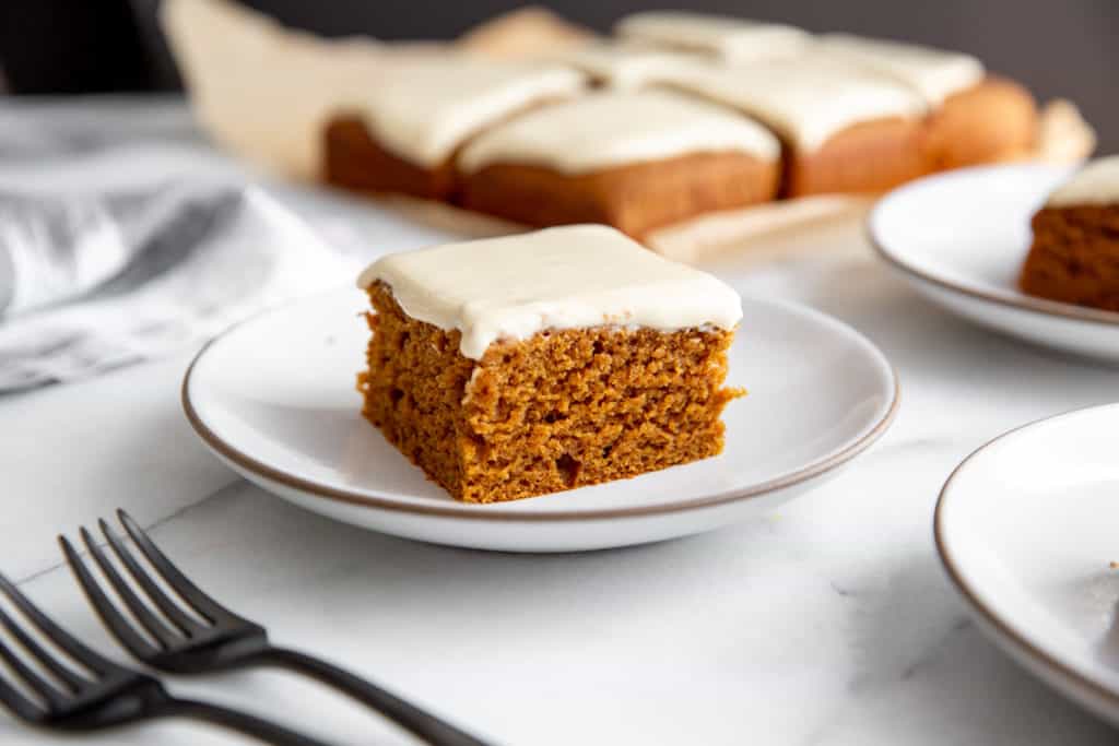 A slice of gingerbread cake on a plate with the rest of the cake and forks in the background.