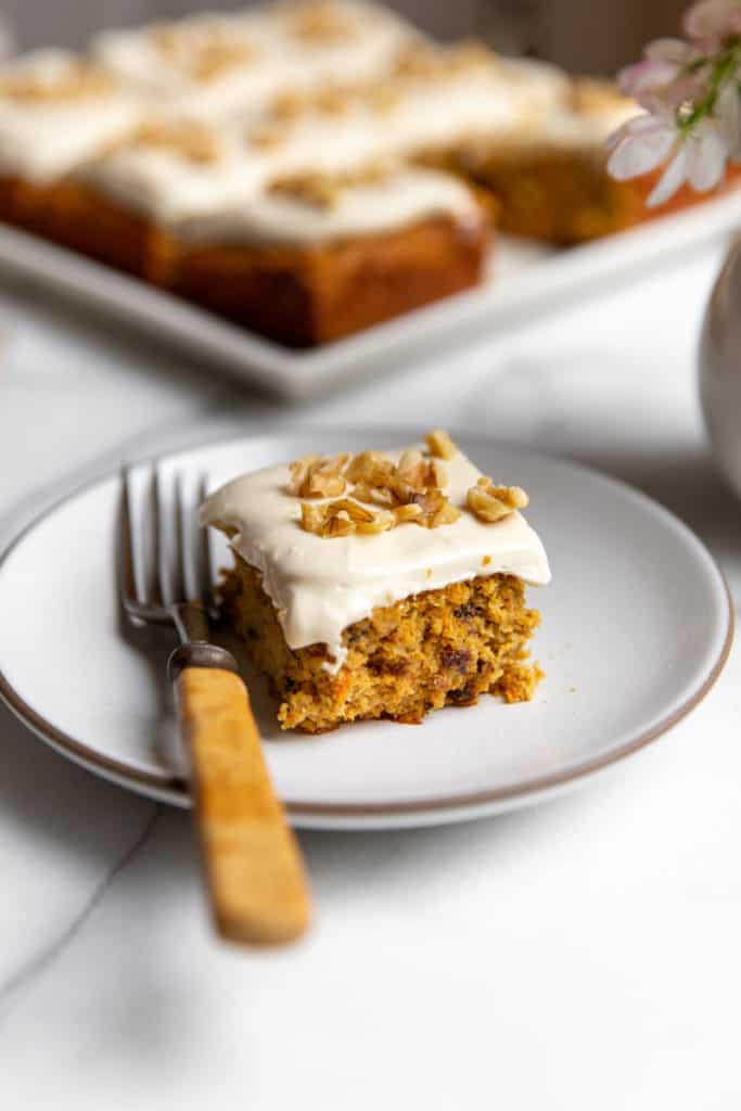 A piece of carrot cake on a plate with a fork, topped with maple cream cheese frosting. 