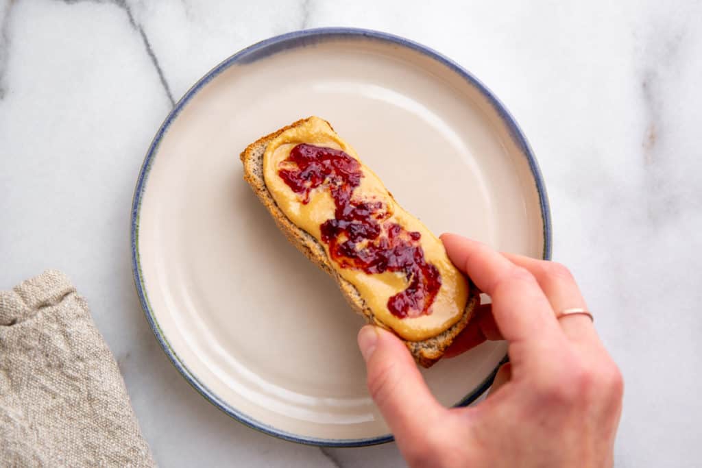 A hand picking up a piece of toasted almond flour bread topped with peanut butter and jelly.