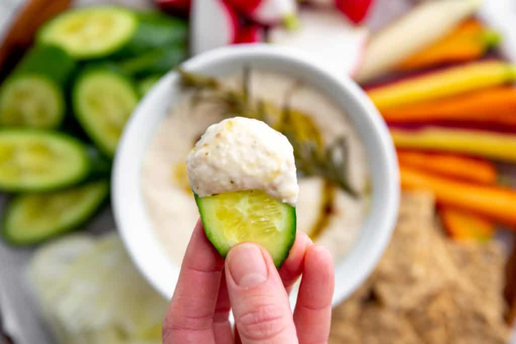 Close-up of a hand holding a cucumber with white bean dip. 
