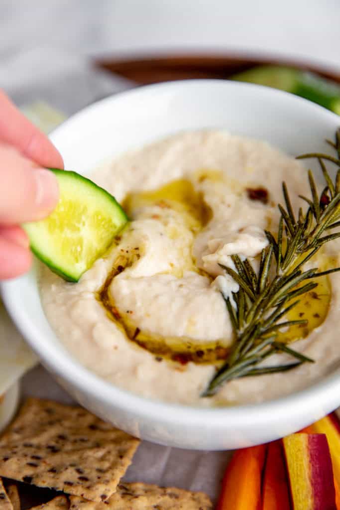 Close-up of a hand dipping a cucumber into a bowl of white bean spread. 