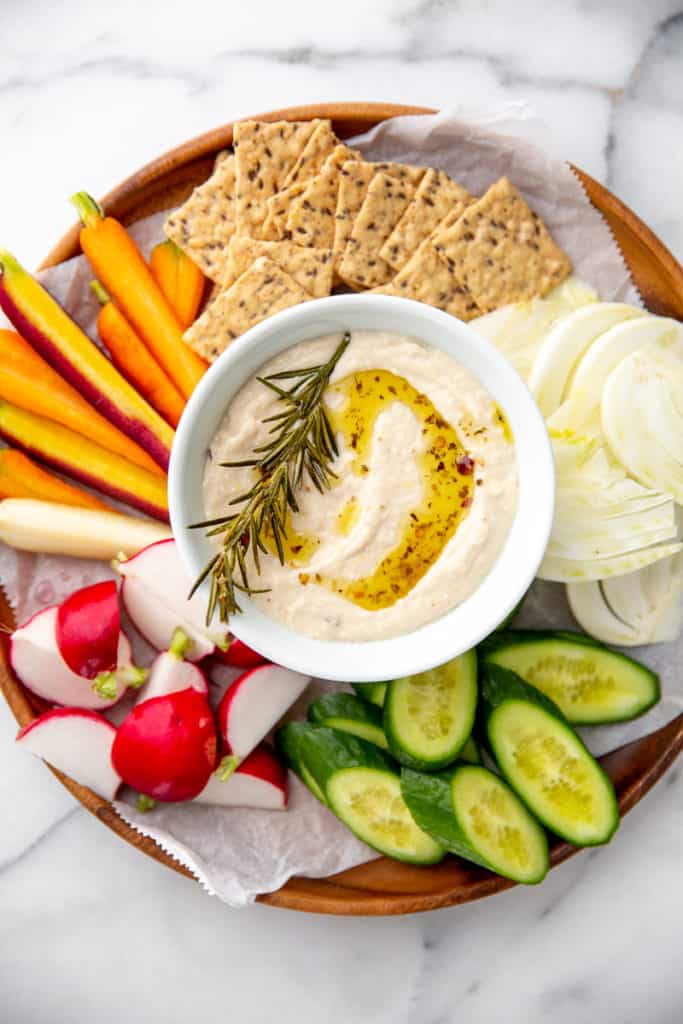 White bean dip in a bowl on a platter surrounded by raw vegetables and crackers for dipping. 
