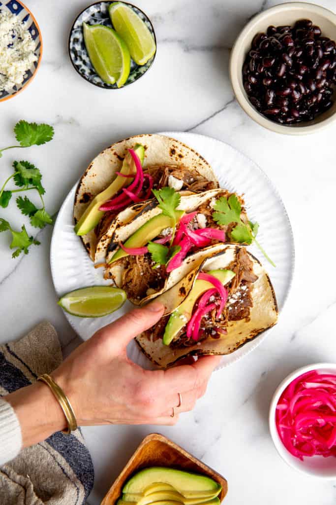 A plate of carnitas tacos topped with quick pickled red onions, with a hand grabbing a taco. 