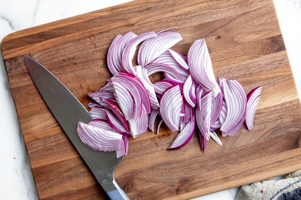 Process shot showing thinly sliced red onions on a cutting board with a knife. 
