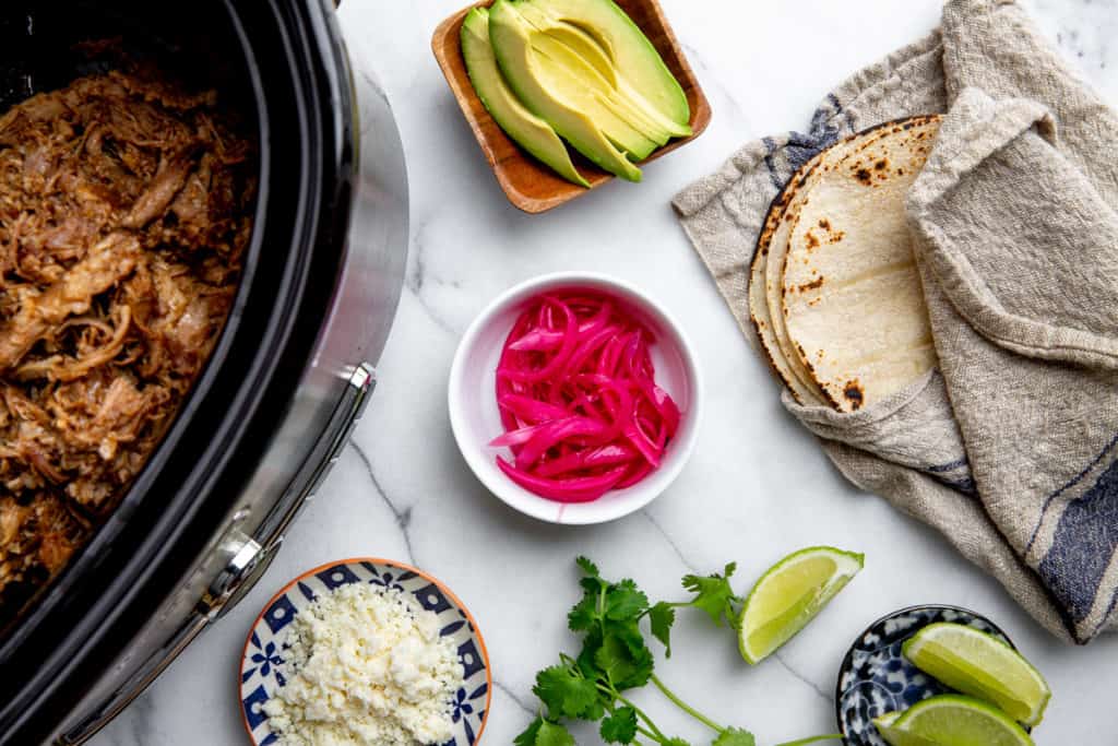 Carnitas in a slow cooker with pickled red onions, avocado slices, tortillas, crumbled cheese and lime wedges alongside. 