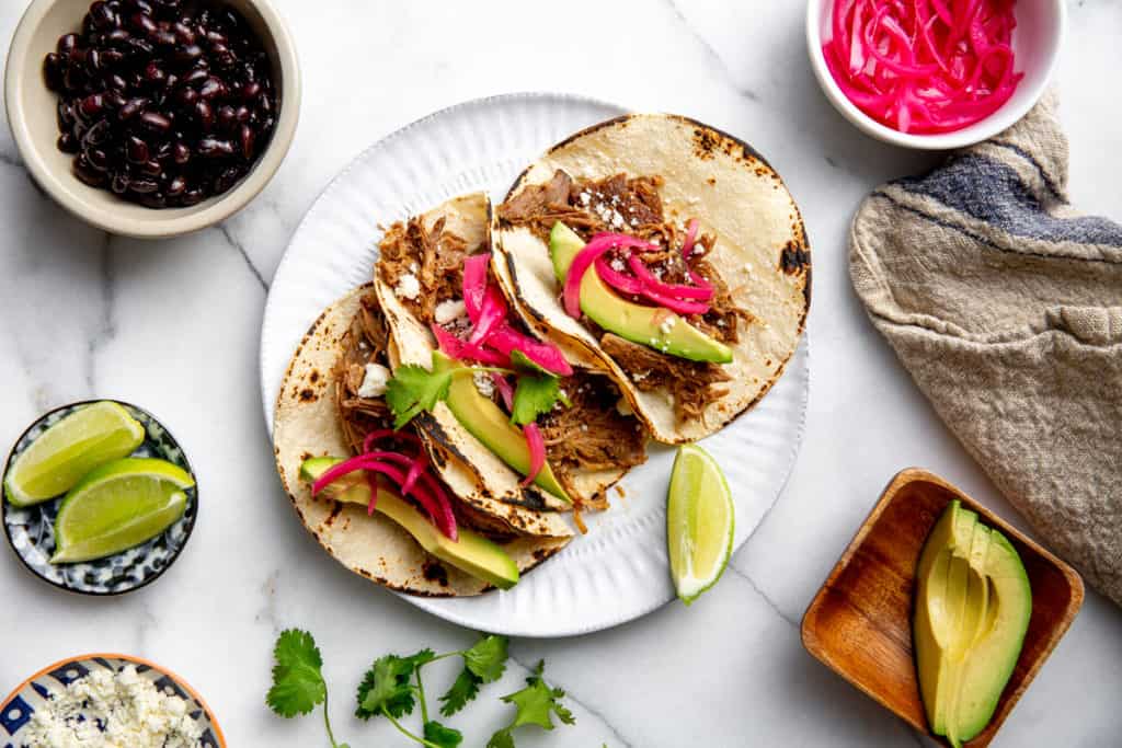 Slow cooker carnitas tacos on a plate topped with avocado slices and quick pickled red onions. 