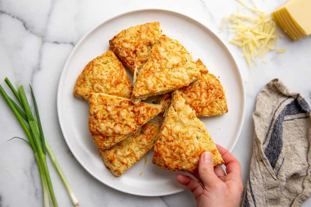 A hand grabbing a cheese scone off of a plate. 