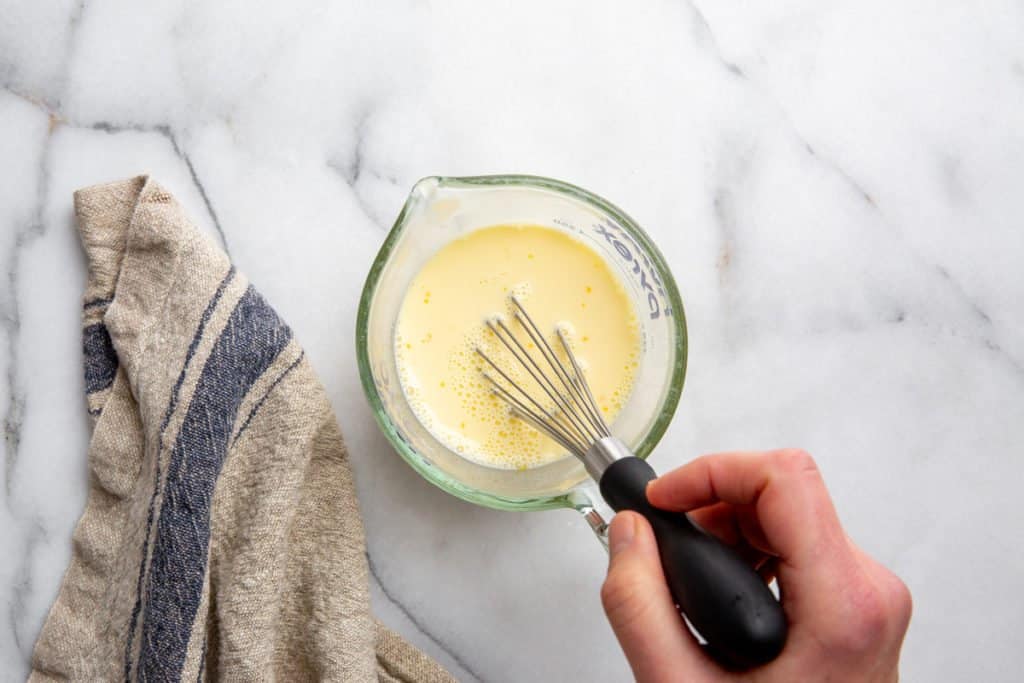 Process shot showing a hand whisking milk and egg in a measuring cup. 