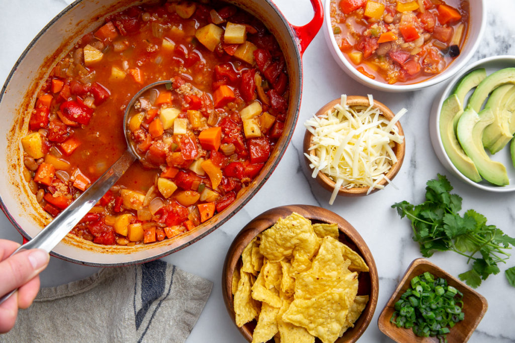 The pot of chili on a countertop surrounded by toppings, including shredded cheese, avocado and tortilla chips. 