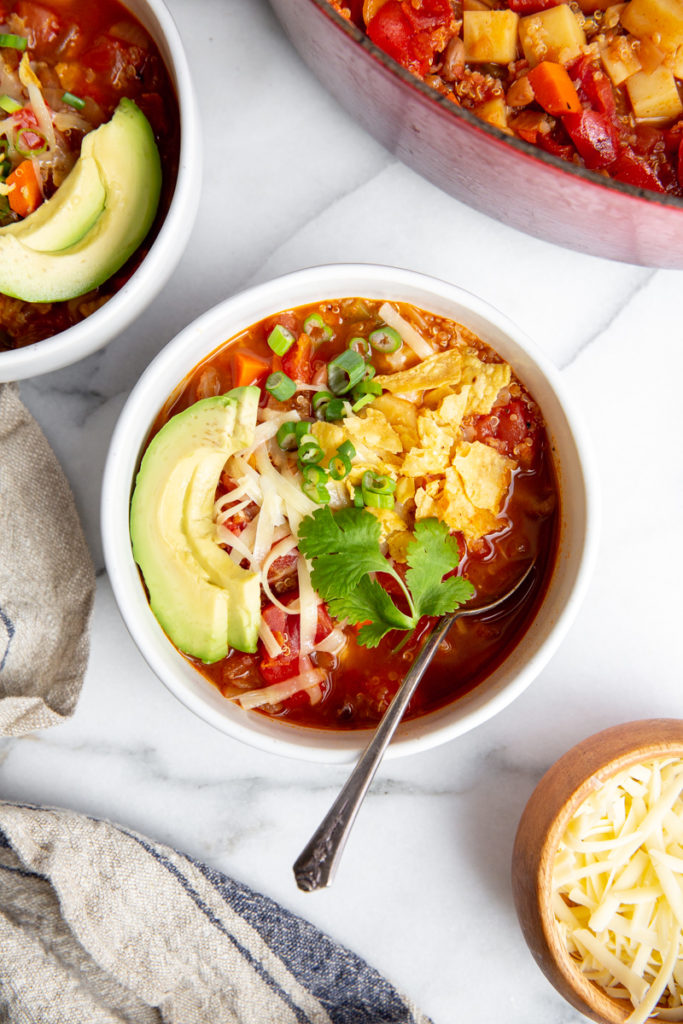 Vegetable quinoa chili in a bowl, topped with cheese, avocado and crushed tortilla chips. 