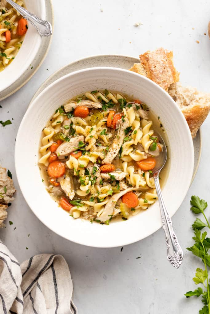 Easy homemade chicken soup from scratch in a bowl with a spoon, with a torn baguette alongside. 
