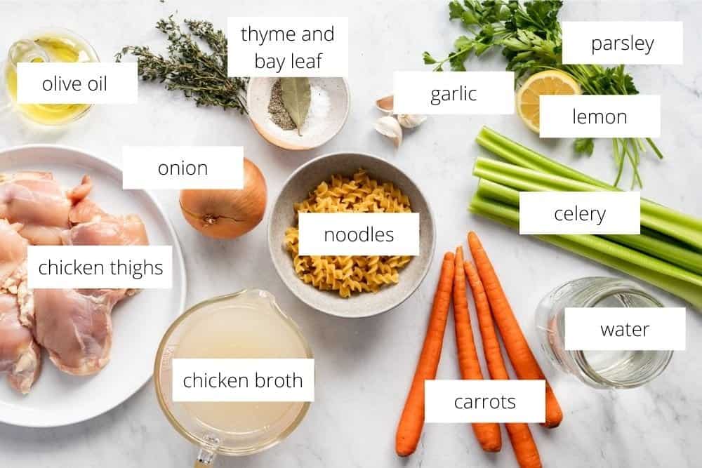 All of the ingredients for the homemade chicken soup recipe, arranged on a white surface with labels. 