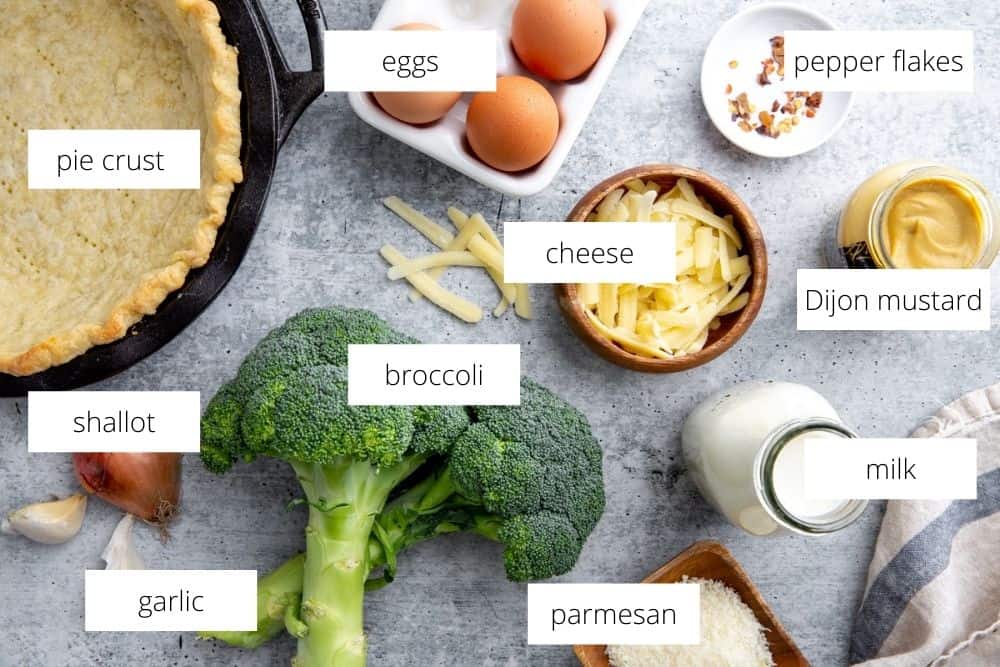 All of the ingredients for the broccoli quiche recipe on a gray surface with labels. 