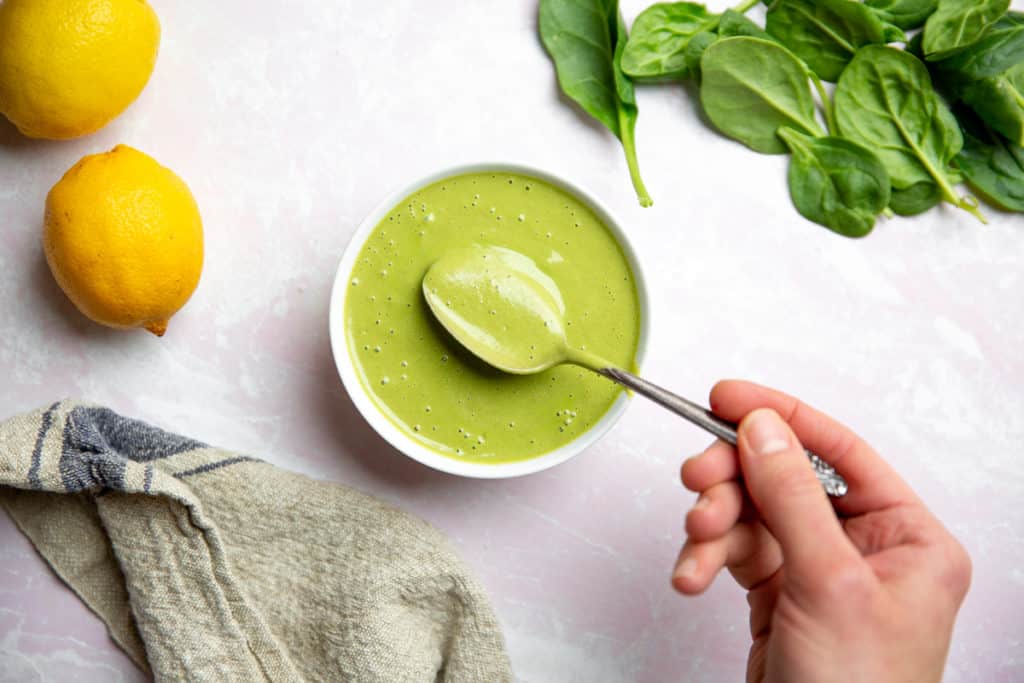 A hand holding a spoon of green tahini sauce in a bowl. 