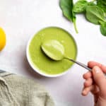 A hand holding a spoon of green tahini sauce in a bowl.