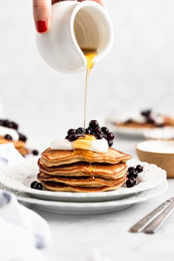 A hand pouring maple syrup on a stack of banana oat pancakes. 