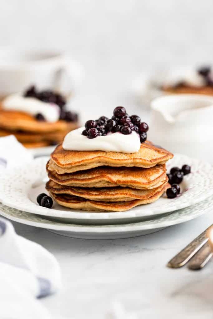 A stack of oat pancakes on a plate topped with yogurt and blueberries. 