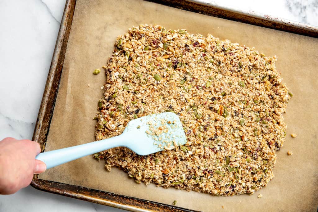 A hand spreading the granola clusters mixture onto a parchment lined baking sheet using a spatula. 