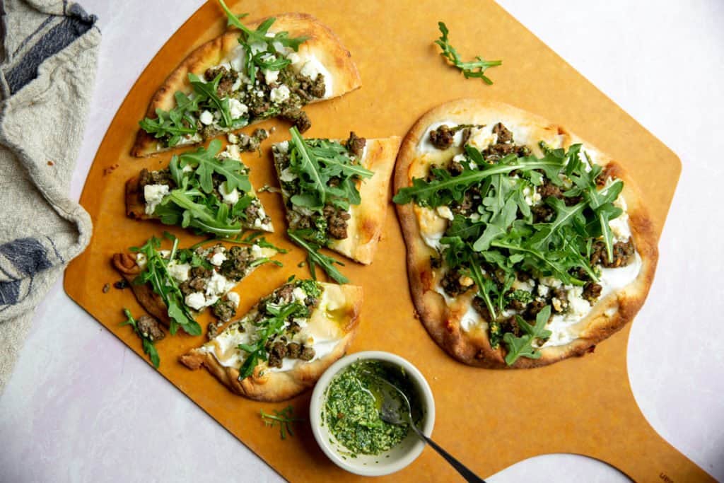 Baked lamb flatbread pizzas topped with baby arugula on a pizza board with mint pesto alongside.
